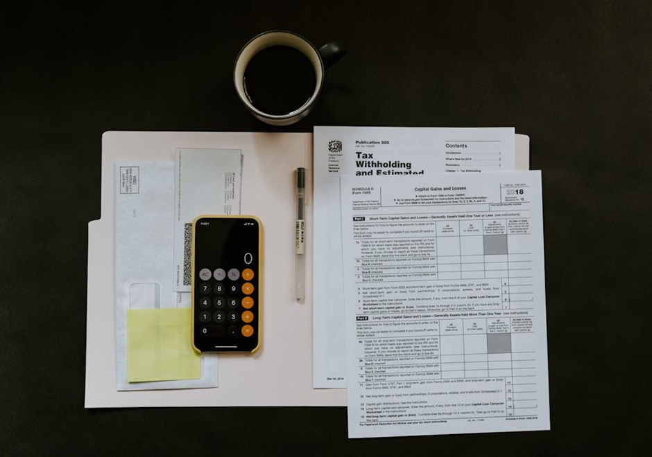 A flat lay of tax preparation materials including IRS tax forms, a smartphone with a calculator app open, a pen, envelopes, sticky notes, and a cup of black coffee on a dark surface.

